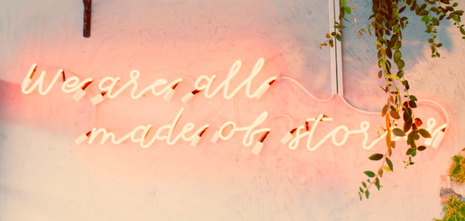 muted colours image of neon sign (under shelf with plants and books on) saying "We are all made of stories"