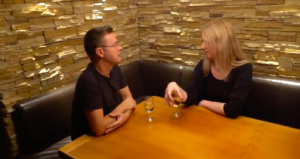 Still from video of Christopher Allen and Helen Rye sitting at a table talking over very small glasses of wine