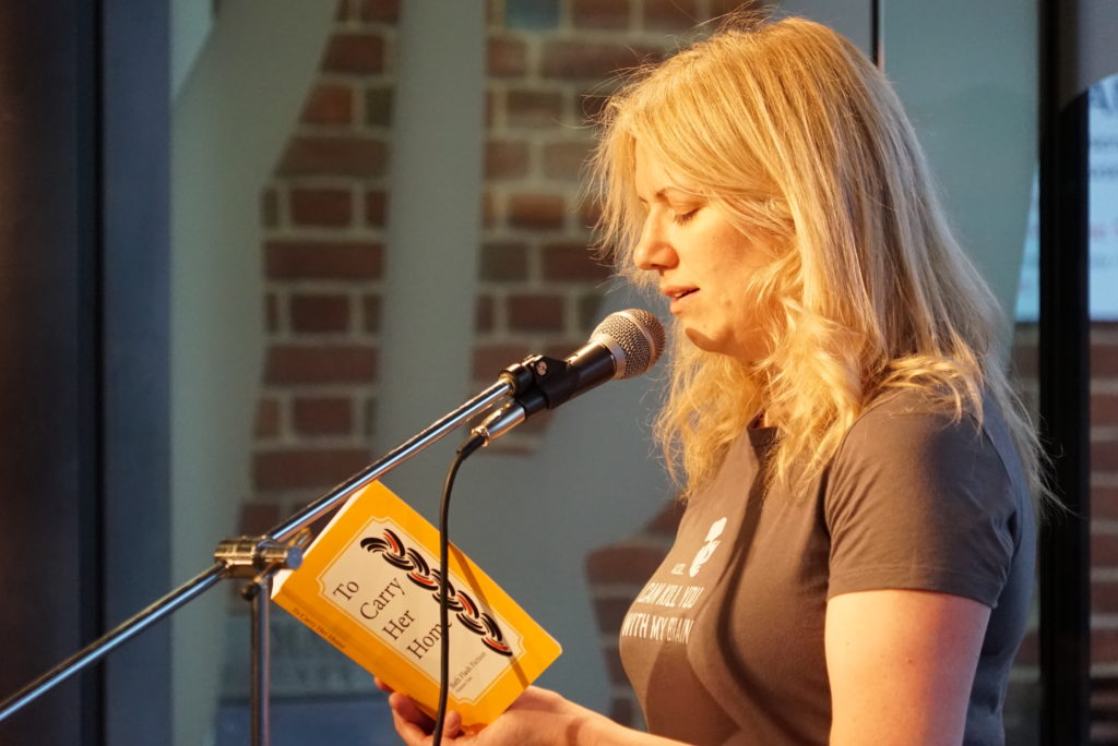 Photograph of Helen Rye reading her flash fiction story One in Twenty-Three from the anthology To Carry Her Home. Helen has shoulder length blonde hair and a grey t-shirt with the Firefly quotation, "Also, I can kill you with my brain". She is speaking into a microphone. The backdrop is the glass, steel and brickwork of Marzano's, The Forum, Norwich